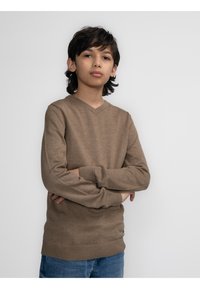 Brauner V-Ausschnitt-Pullover aus weichem Stoff mit langen Ärmeln und geripptem Saum, getragen über blauen Jeans. Das Model steht mit verschränkten Armen.