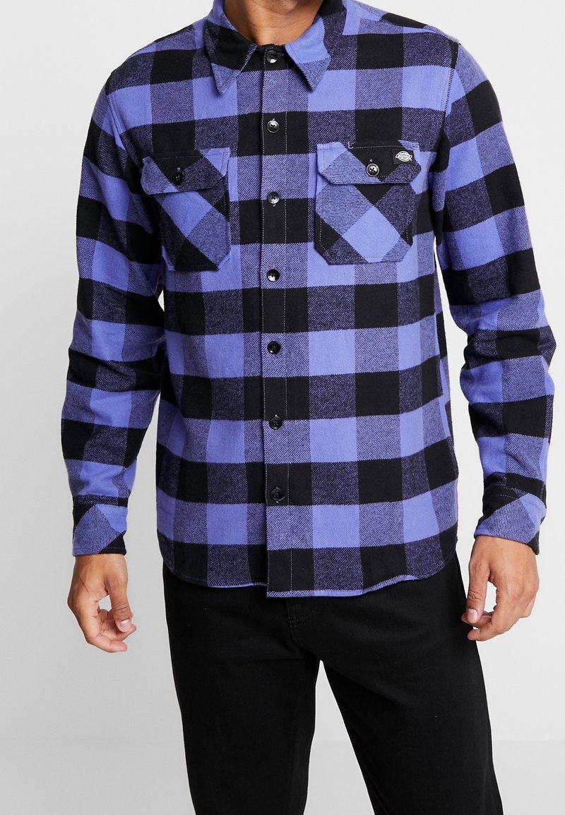 Homme portant une chemise à manches longues à carreaux violets et noirs avec deux poches poitrine à boutons et un pantalon noir.