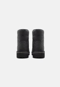 Un paio di stivali neri alla caviglia con suole spesse, cuciture visibili e colli imbottiti, mostrati da dietro su uno sfondo bianco.