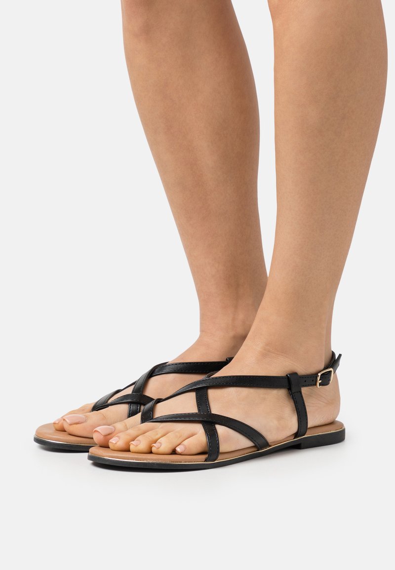Black leather sandals featuring crisscross straps and an ankle buckle. Flat sole with a tan footbed and open-toe design.