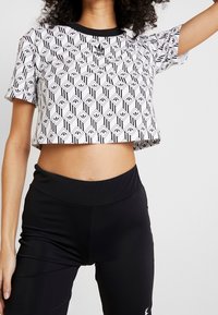 Frau in einem weißen, kurz geschnittenen Adidas-T-Shirt mit einem schwarzen geometrischen Muster und schwarzen, hoch taillierten Leggings, die mit einem erhobenen Arm steht.