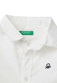 Biała bawełniana koszula z wywijanym kołnierzykiem, z zapięciem na guziki oraz granatowym haftowanym logo po lewej stronie, w pobliżu guzików. Widoczna metka.