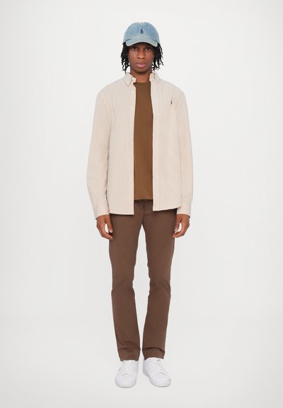 Bežinė ir balta dryžuota marškinėlių priekyje virš rudos t-marškinio, rudos prigludusios kelnės ir baltos sportbačiai, uždėti šviesiai mėlynas kepurė.