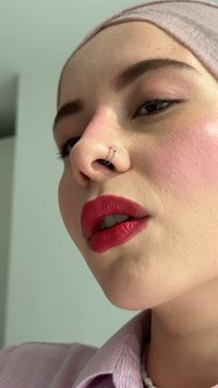 Primo piano di un volto con pelle liscia, una sciarpa rosa chiaro, rossetto rosso audace, un anello nasale a doppio cerchio in argento e accenti di trucco sottili.