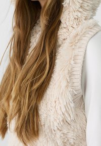 Largo cabello castaño claro sobre un chaleco de felpa color crema, que se lleva sobre una blusa blanca de manga larga, contra un fondo sencillo.