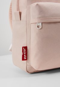 Vaaleanpunainen kangaskassi, jossa on etutasku, valkoinen vetoketju ja punainen Levi's-logo tarrana sivulla. Sujuva pinta ja jämäkkä muotoilu.