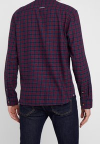 Punainen ja laivastonsininen ruudullinen pitkähiha paita, jossa on napit manseteissa, suora leikkaus, sivuhalkiot ja lenkki niskan takana ripustamista varten.