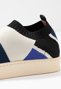 Stickad sneaker i svart, vitt, marinblått och blått med en texturerad ovandel och elastisk muddkant; har en beige gummisul och bakre dragflik.