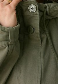 Main tenant le col d'une veste verte boutonnée avec deux boutons visibles et un tissu texturé.