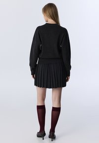 Vrouw staat met haar rug naar de kijker, gekleed in een zwarte sweater, een geplooide zwarte rok, doorschijnende bordeauxkleurige kniehoogsokken en zwarte hakken, tegen een effen achtergrond.