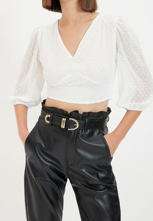 Wit, kortom mouwen, polka dot crop top met een V-hals; gecombineerd met hooglopende, zwarte neplederen broek en een riem met gouden gesp.