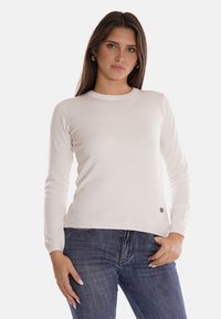 Maglione a manica lunga beige chiaro in morbido tessuto con scollatura rotonda e polsini a coste. Presenta un piccolo logo all'altezza dell'anca. Indossato con jeans scuri.
