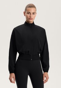 Černá zipová bunda s vysokým límcem, elastickými manžetami a zkráceným lemem. Vyrobena z hladké tkaniny, má strukturovaný design.