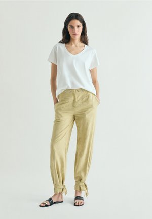 Femme debout sur un fond uni, portant un t-shirt blanc à col en V, un pantalon beige taille haute avec poignets noués, et des sandales noires à enfiler.