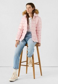 Joven mujer sentada en un taburete de madera, vistiendo una chaqueta acolchada rosa claro con capucha de piel, camiseta blanca, jeans azules y zapatillas altas color crema.