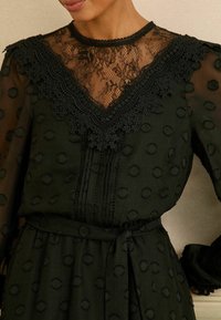 Černé šaty s krajkovým živůtkem s květinovým vzorem, texturovanými puntíky, plisováním a zavázaným pasovým páskem; průhledné dlouhé rukávy s volánkovými okraji.