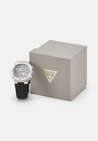 Orologio argentato con quadrante grigio e cinturino in silicone nero, esposto accanto a una scatola grigia texture con logo dorato.