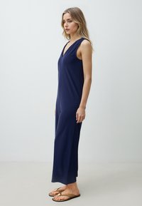 Tuta blu navy con scollatura a V profonda, senza maniche, vestibilità morbida e gambe larghe, realizzata in tessuto morbido e liscio. Indossata con infradito marroni.