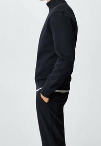 Felpa nera con zip in tessuto morbido, con collo alto e tasche laterali, abbinata a pantaloni scuri. Design semplice e moderno.