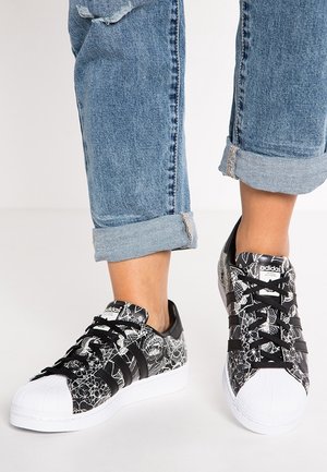 Czarno-białe sneakersy Adidas z kwiatowymi wzorami noszone z podwiniętymi niebieskimi dżinsami na jasnym tle.