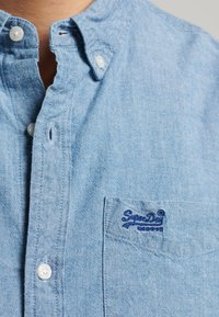 Lichtblauw katoenen overhemd met knopen, voorzien van een kraag, een borstzakje en geborduurde donkerblauwe merknaam op het zakje.