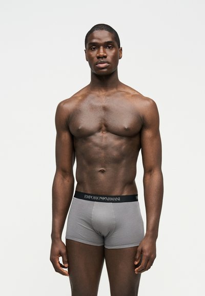 Lihaseline mees seisab, kandmas halli Emporio Armani bokseralusse musta vöökohaga, neutraalne ilme, lihtne hele taust.