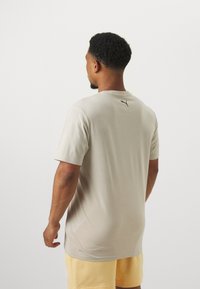 Lys beige kortermet T-skjorte med rund hals og svart logo på baksiden. Laget av mykt stoff, kombinert med gule shorts.
