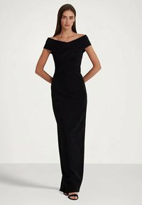 Γυναίκα όρθια με τα χέρια πίσω από την πλάτη, φορώντας ένα μαύρο φόρεμα μέχρι το πάτωμα με ανοιχτούς ώμους και μαύρες γόβες με ανοιχτό δάχτυλο σε απλό φόντο.