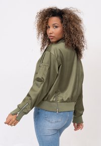 Veste bombardier vert olive avec une finition satinée, dotée de poignets côtelés, de poches latérales zippées et d'une coupe décontractée.