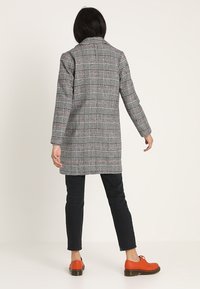 Cappotto a quadri grigio con dettagli rosa, silhouette sagomata, maniche lunghe e spacco posteriore singolo, abbinato a pantaloni neri e scarpe arancioni.