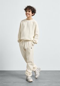 Krämfärgad sweatshirt och joggers i mjukt material, med elastiska band vid ärmar och midja, samt en liten logotyp på byxorna.