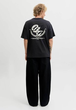 Jeune personne aux cheveux blonds bouclés, debout de dos, portant un t-shirt noir oversize avec un texte graphique blanc et un pantalon large noir.