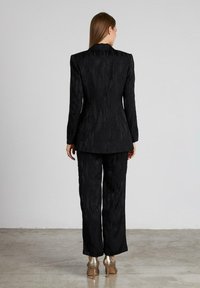 Traje negro a medida con tela texturizada, que presenta una chaqueta estructurada con hombros marcados y pantalones rectos. Tacones dorados visibles.