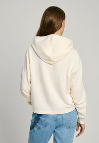 Sudadera color crema con capucha y cordón, hecha de tela suave. Combinada con jeans de un azul claro que presentan un sutil patrón azul en el bolsillo trasero.