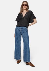 Black top with a deep V-neck and lace trim, paired with wide-leg blue jeans and leopard print shoes. Model wears oversized sunglasses.