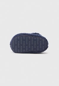Zapatilla de color azul marino con tejido suave y peludo y suela de goma impresa con un patrón de texto repetido que dice "crocs".