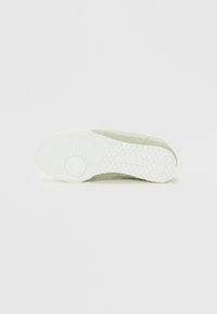 Suela de zapatilla verde claro con suela exterior de goma blanca texturizada que presenta patrones circulares y en zigzag, y la impresión del logo de Lacoste.