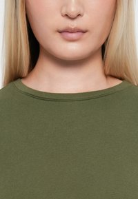 Tricou verde măsliniu, cu guler rotund ribanat, având o textură netedă, cu un design subtil și ajustat și un decolteu ușor brut.