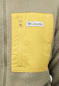 Μπεζ φλις μπουφάν με κίτρινη τσέπη στο στήθος με φερμουάρ, που φέρει το λογότυπο της μάρκας Columbia στην επάνω δεξιά πλευρά της τσέπης.