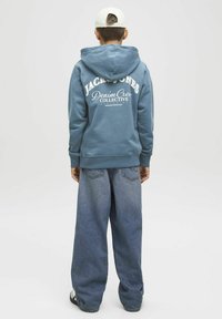 Felpa con cappuccio blu con testo del logo bianco sulla schiena, abbinata a jeans di denim baggy azzurri. Il modello indossa un cappellino bianco e scarpe nere.