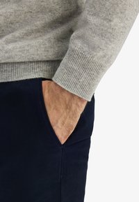 Ljusgrå stickad tröja med ribbad mudd som bärs över mörkblå byxor, vilket framhäver handen i fickan och den fina texturen på tyget.