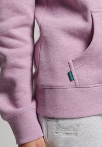 Sudadera de color lila claro con un bolsillo frontal, puños y dobladillo acanalados. Etiqueta con el logotipo de "Superdry" en color teal. Combinada con pantalones de chándal gris claro.