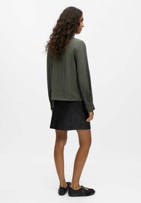 Olivgrünes Langarmhemd aus strukturiertem Stoff, kombiniert mit einem schwarzen Lederrock. Rutschfeste schwarze Schuhe runden das Outfit ab.