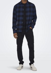 Man som bär en svart och blå rutmönstrad skjorta med knappar över en svart t-shirt, svarta byxor och vita sneakers med blå detaljer.