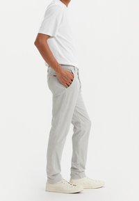 Man wearing light gray pants with hand in pocket, white t-shirt, and off-white sneakers, standing against a plain white background.