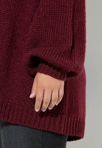 Maglione a maglia color borgogna con superficie texturizzata, polsini e orlo a coste. Mano appoggiata sul maglione, che mostra un tono di pelle liscio.