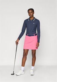 Tengerészkék hosszú ujjú póló fehér gallérral, hozzá illő rózsaszín golf szoknyával. Fehér tornacipő és egy golfütő egészíti ki a megjelenést.