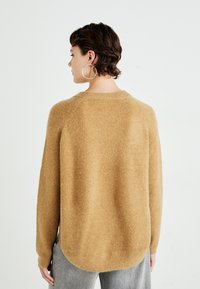 Person with short brown hair wearing large hoop earrings, camel-colored fuzzy sweater, and light gray pants, viewed from behind against white background.