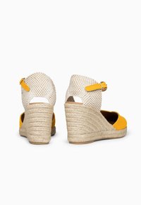 Sandalias alpargatas de cuña con tacones tejidos beige, punteras amarillas y correas al tobillo con hebillas, vistas desde atrás y de lado.