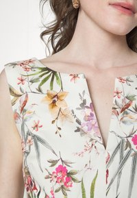 Robe florale présentant un décolleté en V, en tissu léger avec un motif de fleurs roses et jaunes, et de feuilles vertes sur un fond blanc.